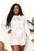 A woman modeling a matching two-piece lounge set featuring a long-sleeve top and shorts in a white and light gray floral jacquard pattern, set against a plain white wall with a plant and chair partially visible.