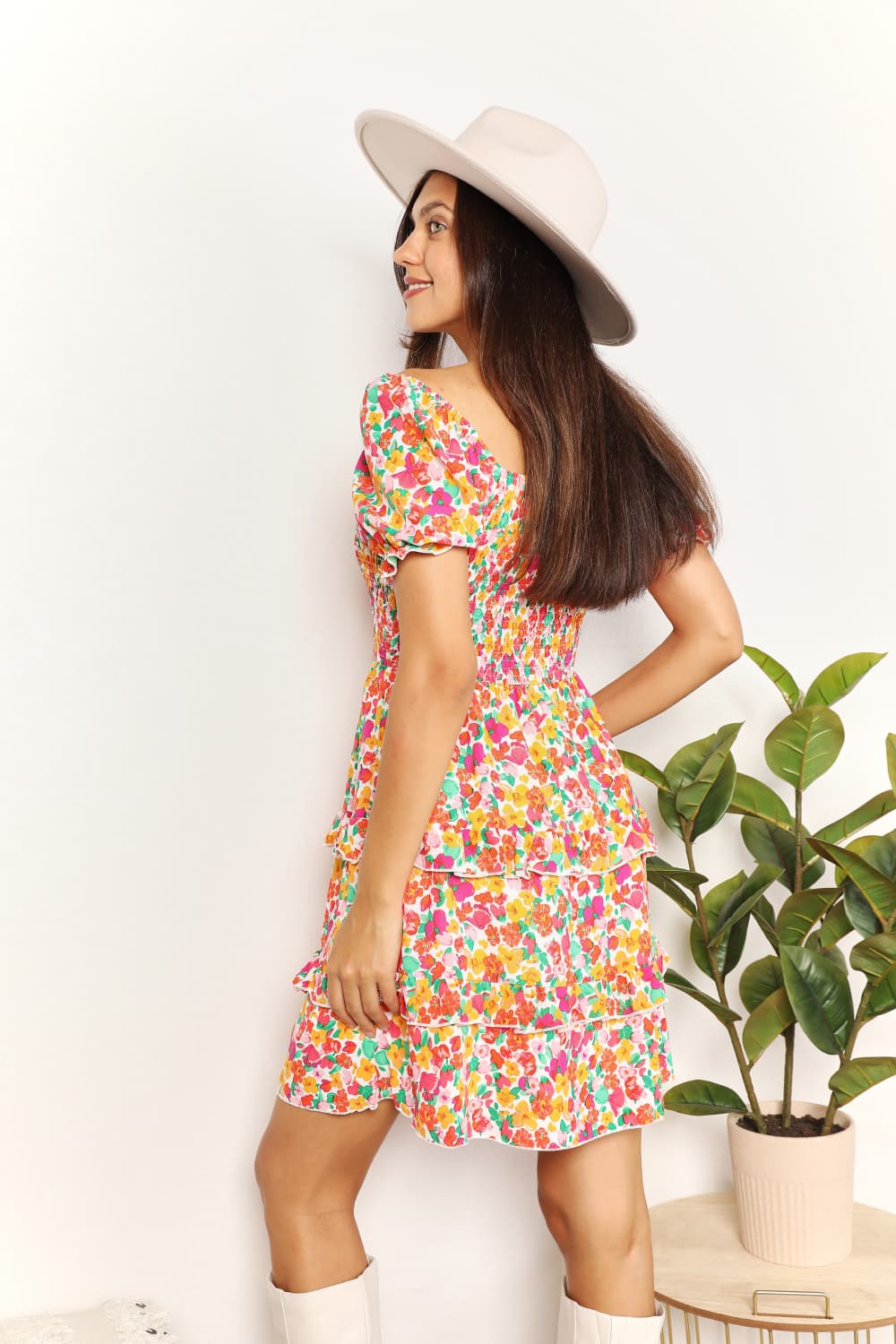 Woman wearing a colorful floral dress and white hat against a white background with a plant.