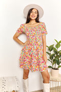 Woman wearing a colorful floral dress with white boots and a hat, standing against a white wall with a plant in the background.