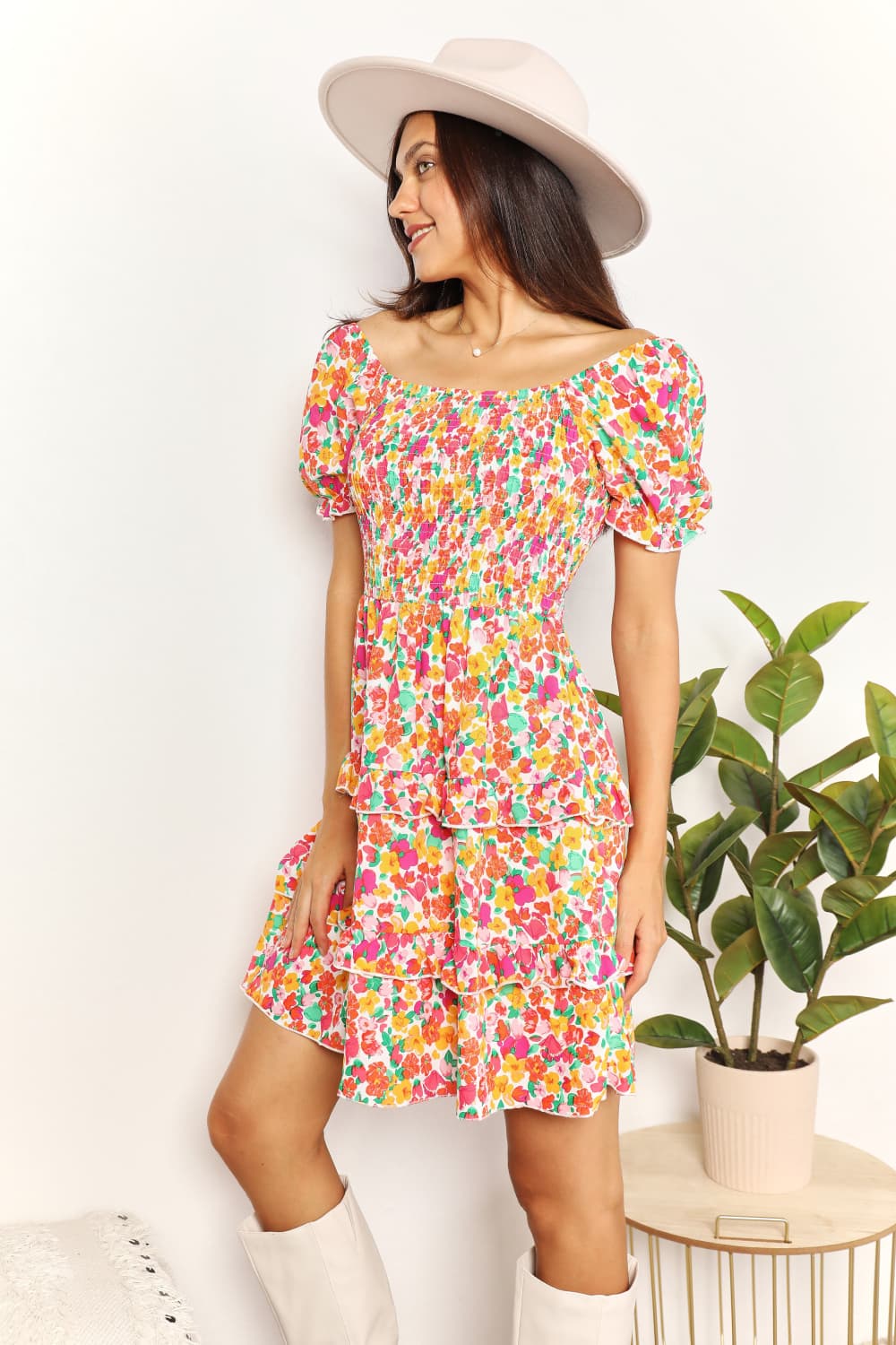 Woman wearing a colorful floral dress with a white hat and knee-high boots, standing against a white wall with a plant in the background.