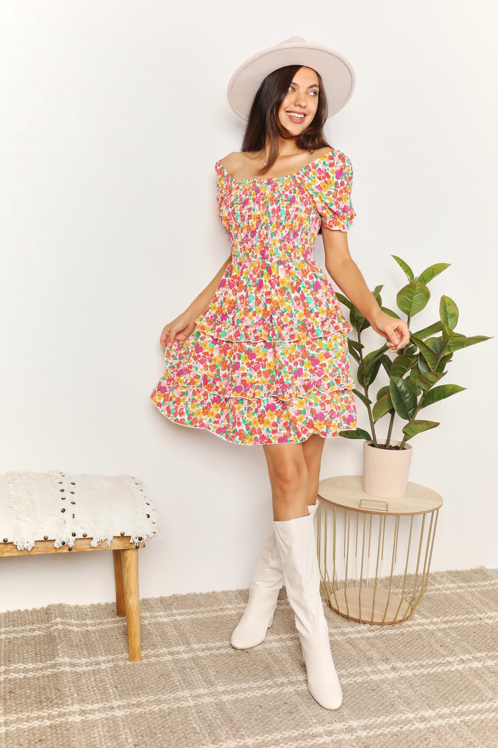 Woman in a colorful floral dress and white boots standing in a room with a plant and small table.
