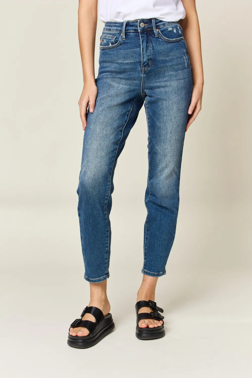 A model wearing a pair of medium wash blue cropped jeans with a tapered leg, styled with black double-strap sandals and a white top against a light beige studio background.